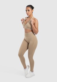 Beige urheilusukka, jossa on syvä V-kaula-aukko ja sen kanssa yhteensopivat korkeavyötäröiset legginsit, jotka on valmistettu joustavasta kankaasta. Valkoiset urheilukengät yksityiskohdilla.