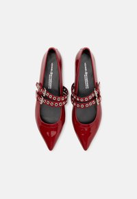 Rote patentlederne High Heels mit spitzen Zehen, ausgestattet mit zwei verstellbaren Riemen, die mit silbernen Ösen verziert sind. Schwarze Innenausstattung.
