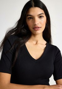 Musta ribbineulottu toppi, jossa V-aukko ja lyhyet hihat, tiukka istuvuus ja sileä pinta. Ei näkyviä kuvioita tai koristeita.