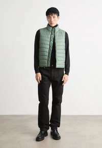 Vesta pufo verde deschis cu cusături orizontale, guler înalt, închidere cu fermoar și logo-ul brodat "HUGO" în roșu, purtată cu pantaloni negri și pantofi.