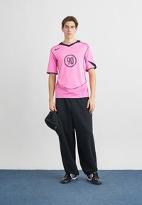 Růžový sportovní dres s krátkými rukávy a černými akcenty, s kulatým logem '90', doplněný o černé široké kalhoty a černou čepici.