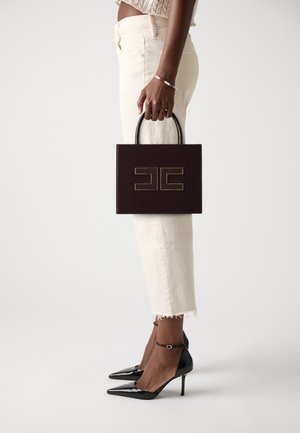 Structured dark burgundy leather handbag featuring a prominent logo, paired with cream-colored ankle-length pants and black heeled shoes.