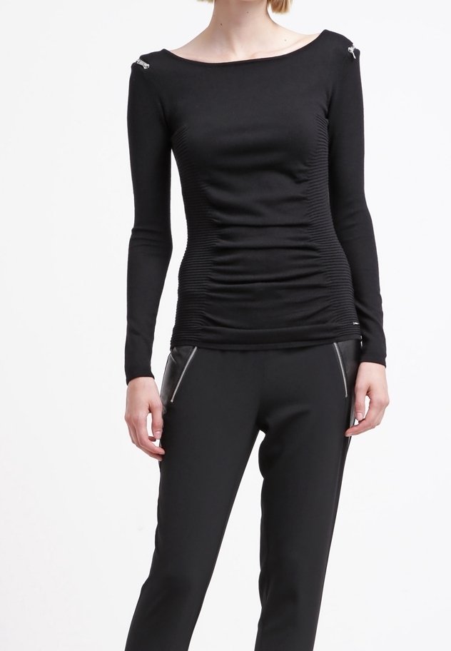 Femme portant un haut noir moulant à manches longues avec des côtés froncés et un pantalon noir slim avec des poches zippées à l'avant, debout devant un fond blanc.