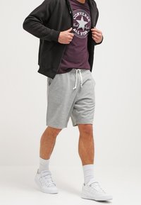 Pantalones cortos de algodón grises con cordón blanco, combinados con una camiseta púrpura con un logo circular de Converse y una sudadera negra con cremallera; zapatillas blancas.