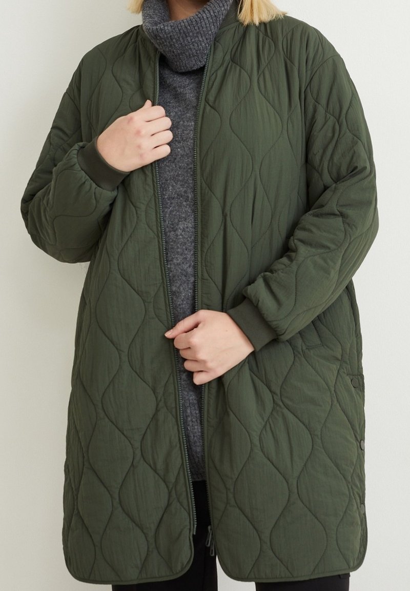 Quilted green jacket with a zip closure, ribbed cuffs, and a textured surface featuring wavy patterns. Worn over a gray turtleneck.