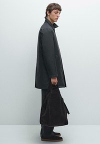 Schwarzer, wasserabweisender Mantel mit hohem Kragen, kombiniert mit einer dunklen Wildleder-Tasche. Das Modell steht vor einem neutralen Hintergrund.