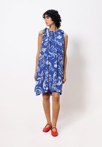 Blaues ärmelloses Kleid mit floralem weißen Muster und gerafftem Ausschnitt. Trägt dazu rote Mary-Jane-Schuhe.