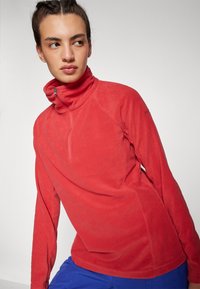Pullover in pile rosso con colletto alto e zip a un quarto. Presenta maniche lunghe e una texture morbida. Decorato con logo sulla spalla.