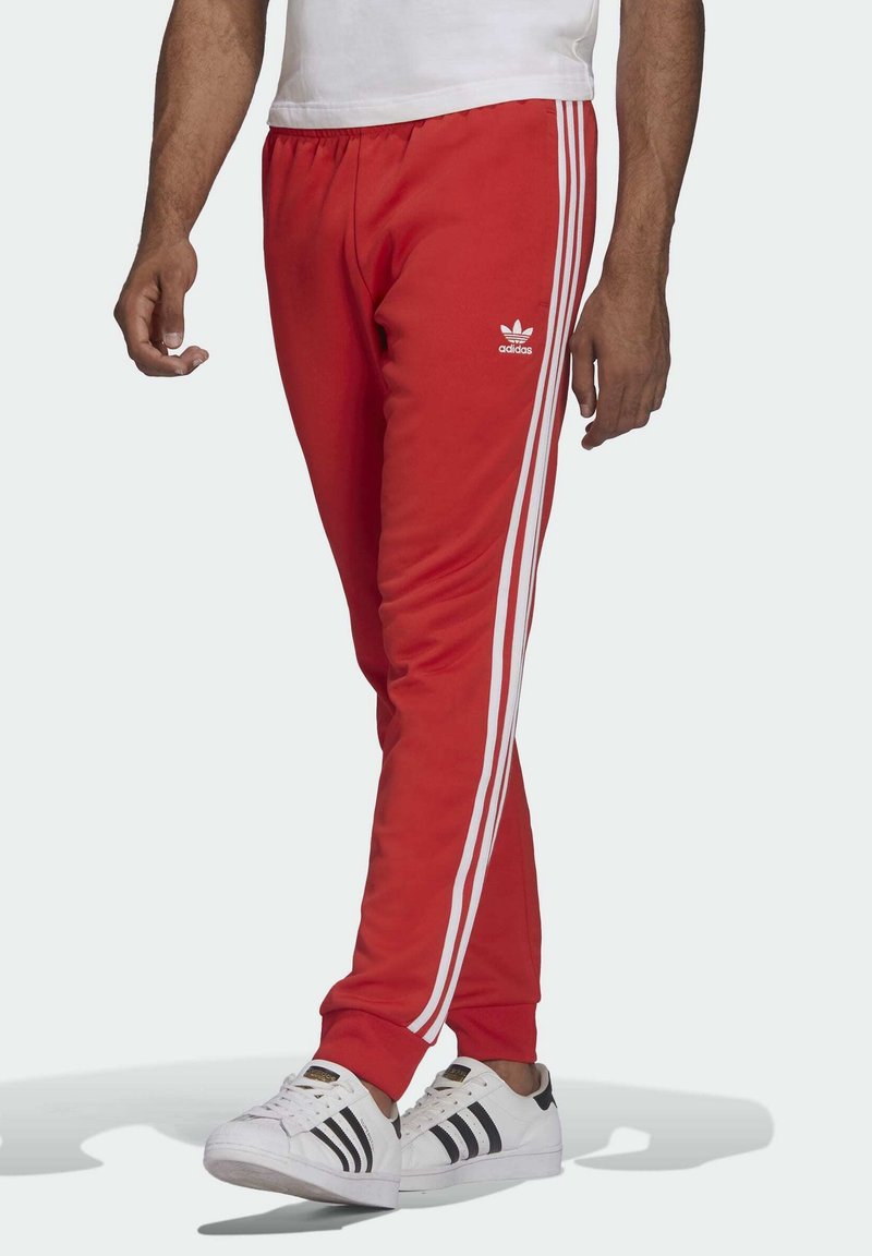 Pantaloni sportivi rossi con design a tre righe bianche, vita elastica e tasche, abbinati a sneakers bianche con accenti neri.