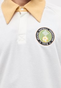 Λευκό ριγωτό πουκάμισο με μπεζ γιακά και στρογγυλή ένθετη ετικέτα της Ποδοσφαιρικής Ομοσπονδίας Heathhe 1976 που εμφανίζει μια μπάλα ποδοσφαίρου στο στήθος.