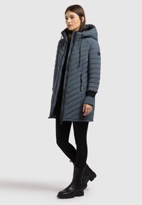 Zateplená sivá parka s kapucňou, diagonálny chevronový vzor, predný zips a bočné vrecká. Nosená cez čierne legíny a topánky.