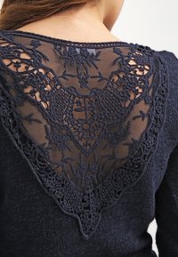 Top azul marino con una tela de punto texturizada. La parte posterior tiene una superposición de encaje detallada en forma de V con patrones florales y bordes en forma de concha.