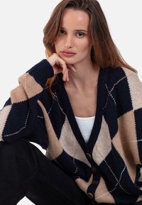 Jeune femme aux longs cheveux bruns, portant un cardigan à losanges bleu marine, beige et crème, reposant son menton sur sa main, assise devant un fond blanc.