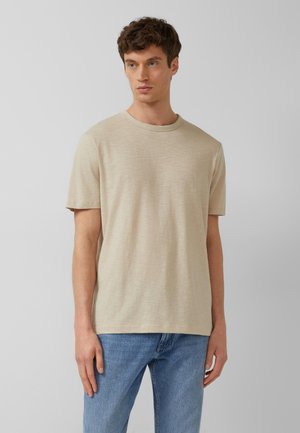 Młody mężczyzna w beżowej koszulce z krótkim rękawem i okrągłym dekoltem oraz jasnoniebieskich dżinsach, stojący na jednolitym jasnoszarym tle.