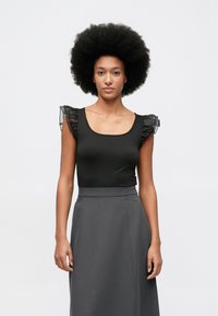 Kobieta z dużą fryzurą afro, ubrana w bezrękawnik czarny z przezroczystymi, falbanowymi ramionami oraz spódnicę w ciemnoszarym kolorze z wysokim stanem.