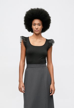 Moteris su dideliu afro šukuosena, vilkinti be rankovių juodą topą su permatomais volanų pečiais ir aukštu juosmeniu tamsiai pilkomis sijonomis.