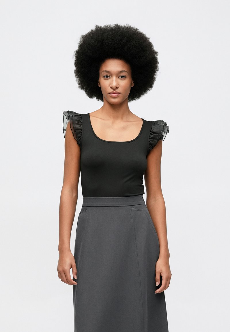 Kobieta z dużą fryzurą afro, ubrana w bezrękawnik czarny z przezroczystymi, falbanowymi ramionami oraz spódnicę w ciemnoszarym kolorze z wysokim stanem.