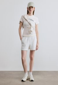 Ανοιχτό μπεζ μπλουζάκι με το κείμενο "Tommy Jeans", λευκά σορτς και αθλητικά παπούτσια. Ένα ανοιχτόχρωμο καπέλο και ένα ανοιχτό μαντίλι τυλιγμένο γύρω από τον ώμο.