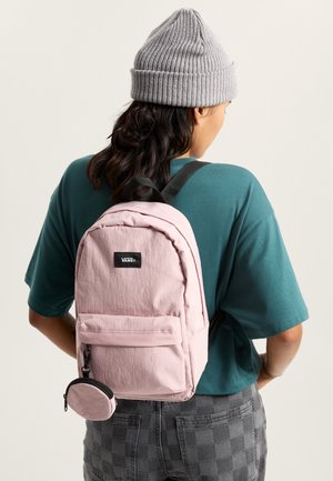 Mochila de color rosa claro con superficie texturizada, bolsillo frontal y un pequeño bolso removible. Llevada por una persona con camisa verde azulado y gorro gris.