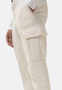 Mann trägt beige Cargo-Hosen mit Klappentasche am Oberschenkel und Hand in der Seitentasche vor weißem Hintergrund.