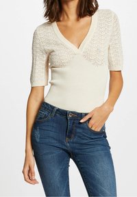 Kortärmad, krämfärgad stickad topp med V-ringning, prydd med strukturerade mönster på axlarna och kroppen. Kombinerad med mörkblå jeans.