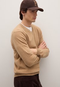 Beiger Pullover mit V-Ausschnitt aus weichem Strickmaterial, kombiniert mit einer braunen Mütze mit Logo. Das Model steht mit verschränkten Armen vor einem einfarbigen Hintergrund.
