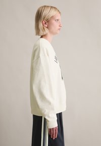 Sweter w kolorze kremowym o luźnym kroju, z ściągaczami na mankietach i grafiką z przodu. W połączeniu z granatowymi spodniami z bocznymi paskami.