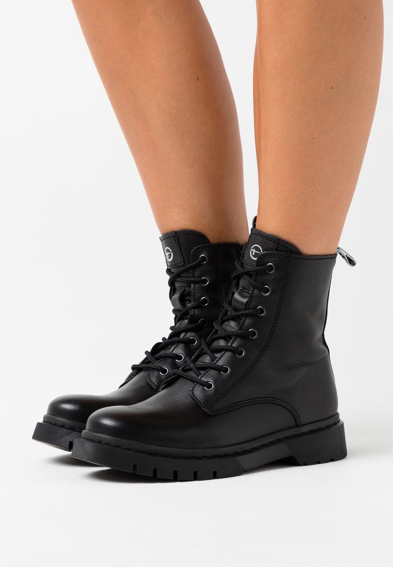 Bottes de combat noires à lacets portées sur des jambes nues, avec une semelle épaisse et une petite languette à l'arrière, sur fond blanc.