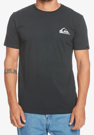 Mann trägt ein einfaches dunkelgraues Kurzarm-T-Shirt mit kleinem weißem Logo auf der linken Brust und hellblauen Jeans.