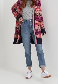 Stickad cardigan med färgglada geometriska mönster i rosa, lila och marinblått, kombinerad med ljusblå jeans och vita sneakers.