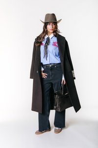 Lange bruine jas over een blauw gestreept shirt met rode borduursels, zwarte high-waisted jeans, bruine schoenen en een zwarte handtas.