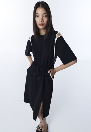 Młoda kobieta stoi z rękami w kieszeniach, ubrana w czarną sukienkę midi z białym falowanym wykończeniem, wycięciami na ramionach i bocznym rozcięciem, w zestawie z czarnymi butami.