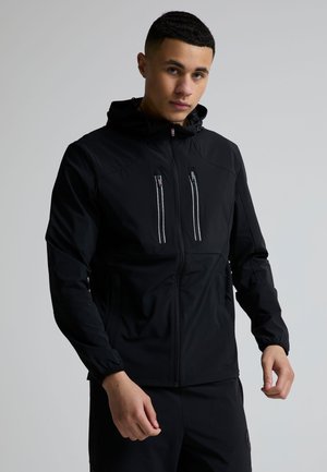 Ung mand iført en sort hættetrøje med lynlåslommer på brystet, stående mod en ensartet lysegrå baggrund.