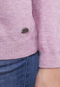 Light pink knitted sweater with ribbed cuffs, featuring a small silver logo patch on the lower side and soft texture. Paired with blue jeans.