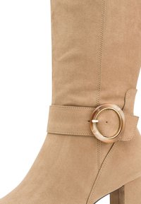 Beige suede ankle boot with a chunky heel, featuring a wide belt strap and a large decorative circular buckle in a marbled finish.