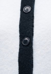 Cardigan noir et blanc présentant une bande centrale noire avec deux boutons noirs, fabriqué en tissu tricoté doux et texturé.
