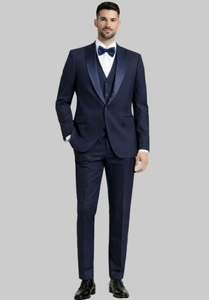 Homme debout, main dans la poche, portant un smoking bleu marine avec des revers en satin, une chemise blanche, un nœud papillon bleu et des chaussures de ville noires.