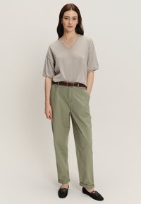 Vrouw staat in een beige kortmouw V-hals top, olijfgroene hoog-getailleerde broek met een bruine riem en zwarte veterschoenen.