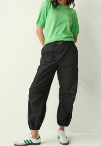 Zwarte cargobroek met een elastische tailleband, taps toelopende boorden en zijzakken, gecombineerd met een groen gebreid T-shirt met korte mouwen en witte sneakers.