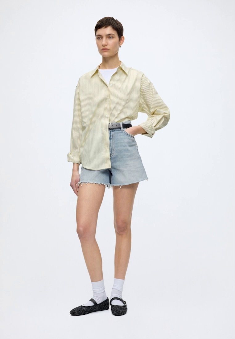 Femme debout portant une chemise oversize rayée jaune pâle, un short en jean clair aux ourlets effilochés, des chaussettes blanches et des ballerines noires à brides.