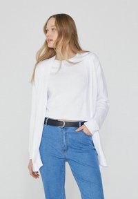 Un cardigan blanc superposé sur un top blanc, associé à un jean bleu. Le cardigan présente une coupe ouverte à l'avant et des manches longues, avec une texture côtelée.