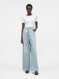 Donna che indossa una t-shirt bianca a maniche corte e jeans a vita alta e gamba larga azzurri chiaro, con una mano in tasca e sandali neri.