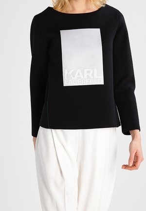Kobieta ubrana w czarną bluzkę z długim rękawem z dużym białym prostokątnym logo z napisem "KARL LAGERFELD" oraz w białe spodnie.