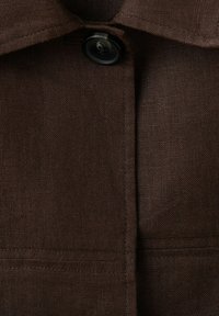 Tissu marron foncé avec des coutures visibles, un bouton noir attaché près du col, mettant en valeur la texture et les détails du vêtement.