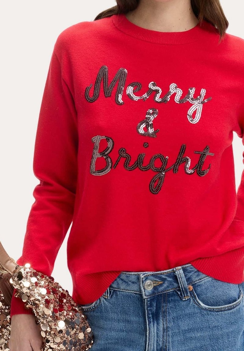 Pull rouge avec texte en sequins noirs et blancs "Merry & Bright", porté avec un jean bleu et un sac à main avec de grandes paillettes brillantes.