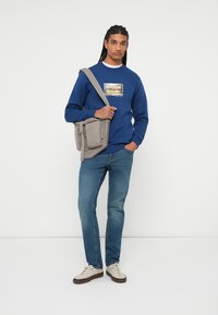 Grijze schoudertas met voorzakken, gedragen met een blauwe sweatshirt met een ingelijste grafische afbeelding en blauwe spijkerbroek, gecombineerd met crèmekleurige sneakers.