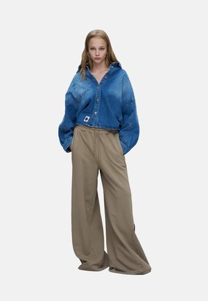 Jeune femme debout portant une veste en jean bleue oversize et un pantalon large beige ample sur un fond uni.