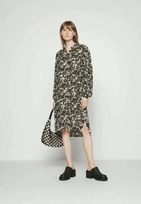 Vestido de mangas compridas, com botões, em preto com padrão floral sobre um fundo bege. Apresenta fendas laterais e um corte descontraído. Acompanhado por uma bolsa de malha e tamancos.