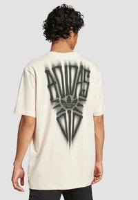 Krémové oversized tričko s velkým černým nápisem "ADIDAS" a grafickými akcenty na zadní straně. Krátké rukávy, měkký textilní povrch.