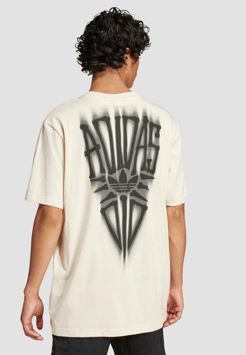 Krémové oversized tričko s velkým černým nápisem "ADIDAS" a grafickými akcenty na zadní straně. Krátké rukávy, měkký textilní povrch.